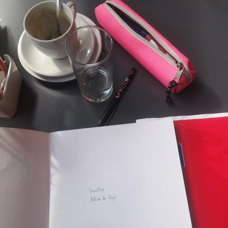 mesa de bar, detalle con taza, vaso, cartuchera y un cuaderno abierto en una hoja donde se lee "Iniciáticos, bitácora de viaje"