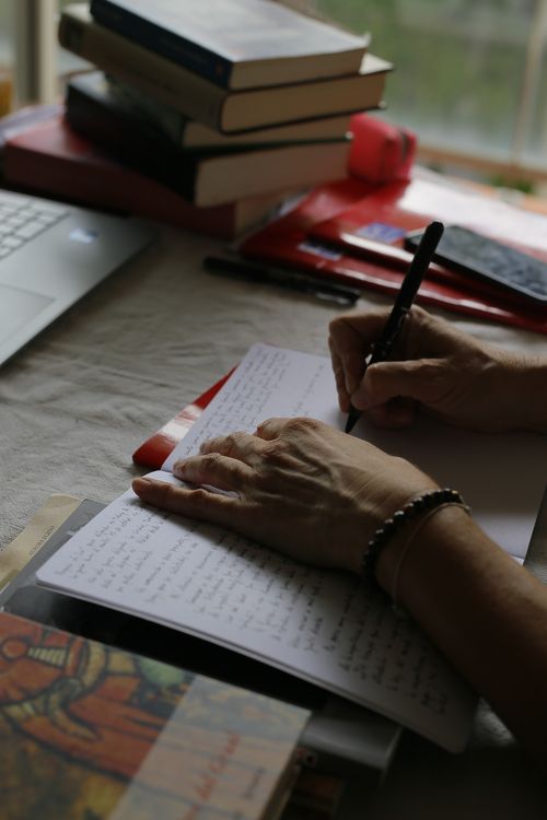 En un escritorio, una mano escribe en un cuaderno, la otra sostiena la hoja. Más allá unos libros y una ventana desde donde viene la luz.