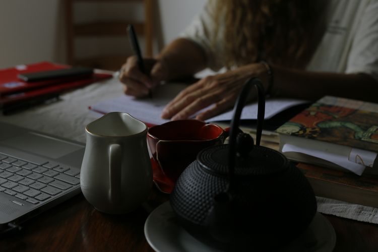 En un escritorio, una mano escribe en un cuaderno, la otra sostiena la hoja. Hay una notebook. Más adelante, una tetera, taza y un jarrito quizás con leche.