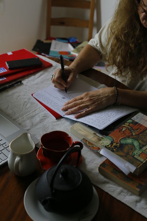 En un escritorio, una mano escribe en un cuaderno, la otra sostiena la hoja. En esta imagen se ve algo más de la persona, una parte de la cara con lentes. Hay una notebook. Más adelante, una tetera, taza y un jarrito quizás con leche.