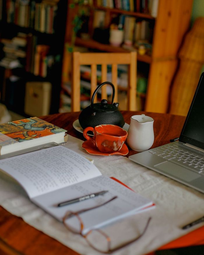 Una mesa con una tetera, taza, jarra, un cuaderno abierto con una lapicera y lentes, un libro cerrado y una notebook. Más lejos hay una biblioteca y una silla.